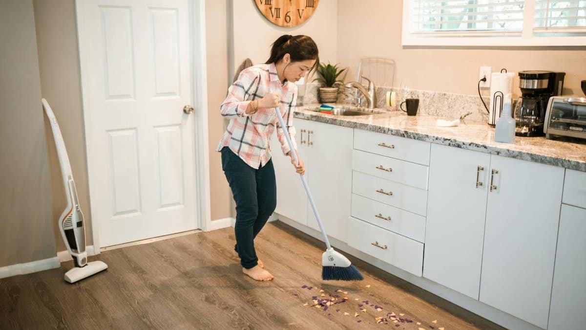 Asian woman sweeping kitchen floor with a broom, performing house chores in a modern kitchen.