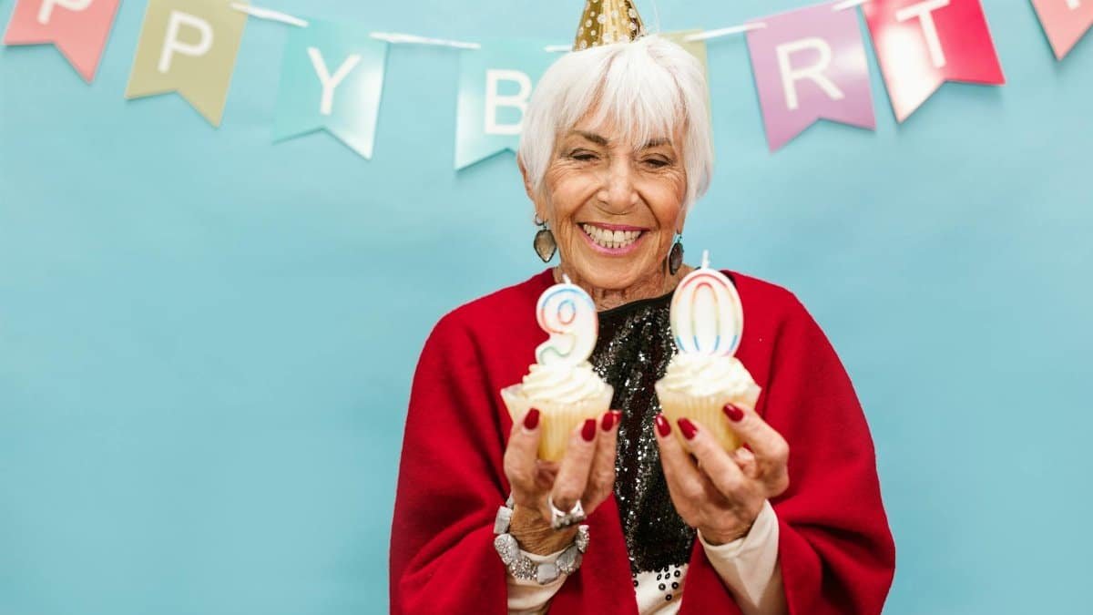 Smiling elderly woman holding cupcakes with candles on 90th birthday party.