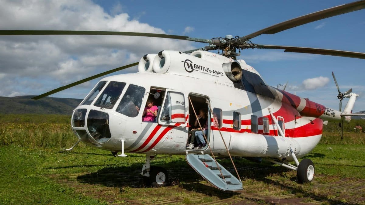 White and red helicopter parked on a grassy field in Kamchatka, Russia under a blue sky.
