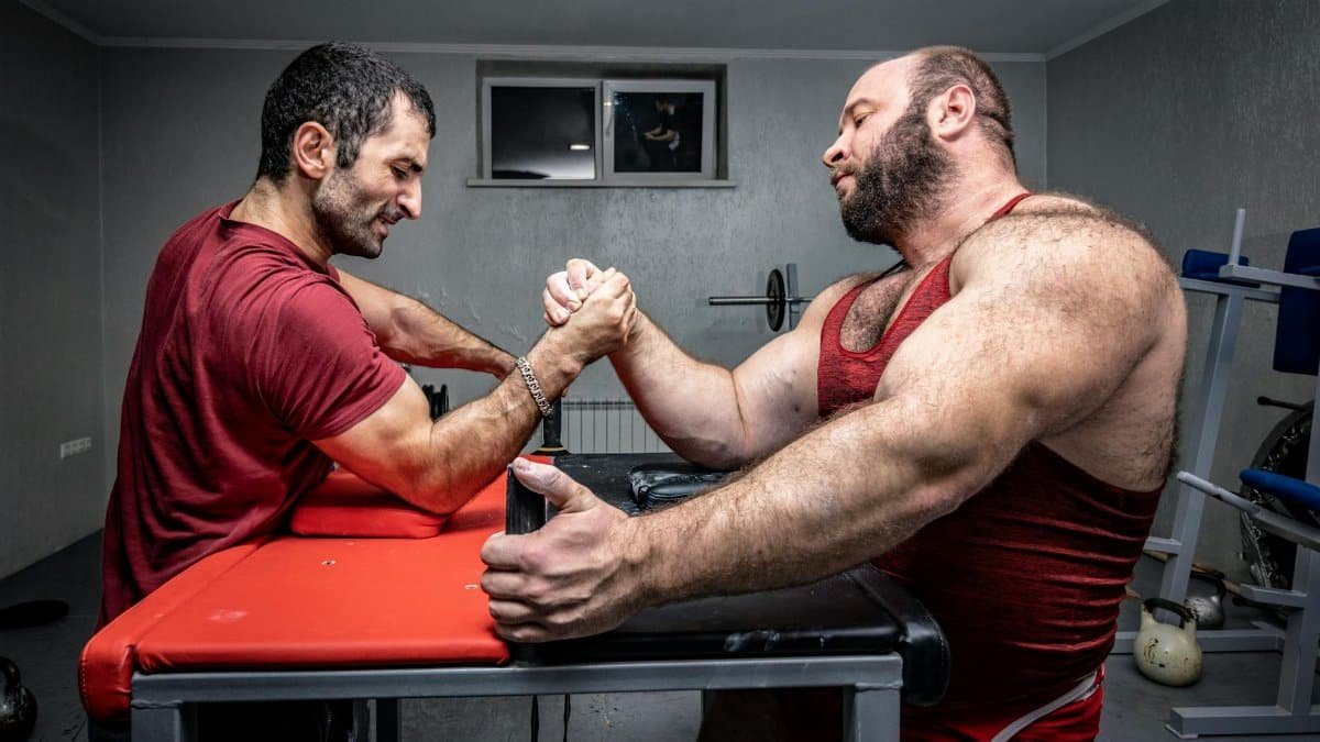 Two muscular men engage in a competitive arm wrestling match in a gym environment.