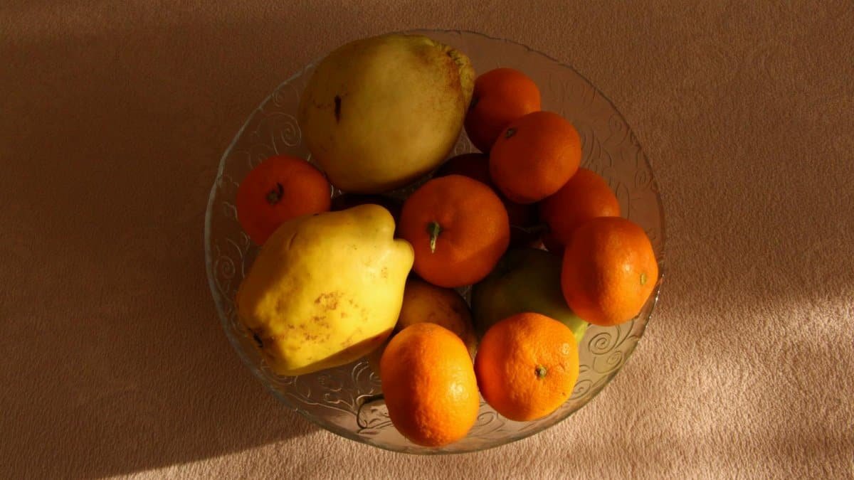 A variety of fresh fruits including quinces and oranges in a glass bowl under warm lighting.