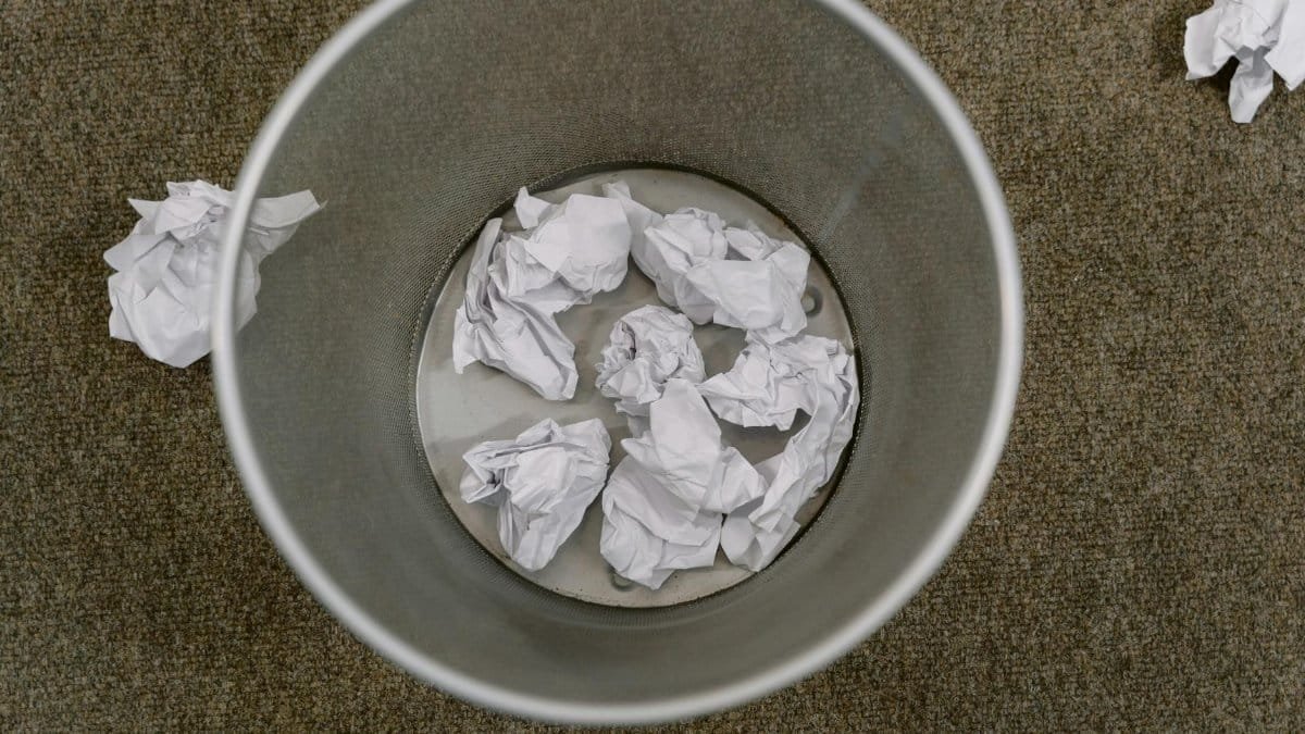 Overhead view of a trash can filled with crumpled paper on a carpeted floor.