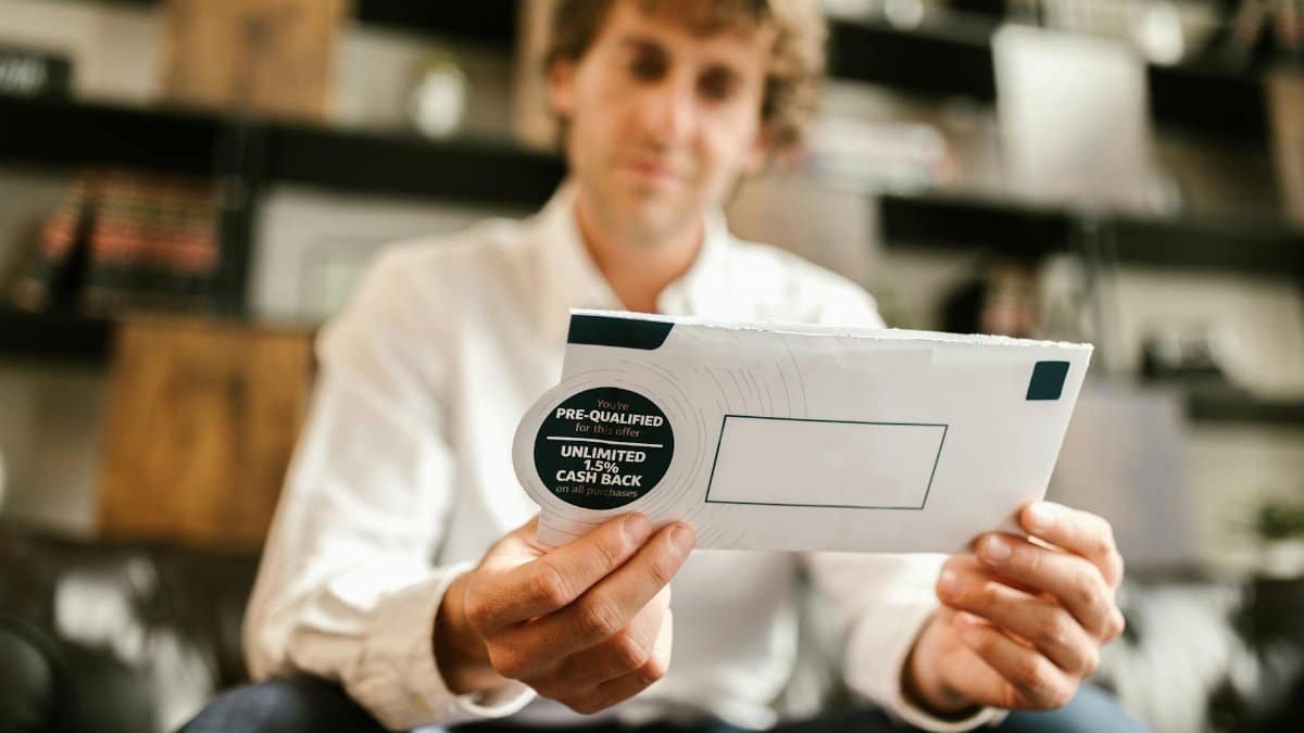 A man sitting indoors, reading an envelope with a cash back offer, blurred background.
