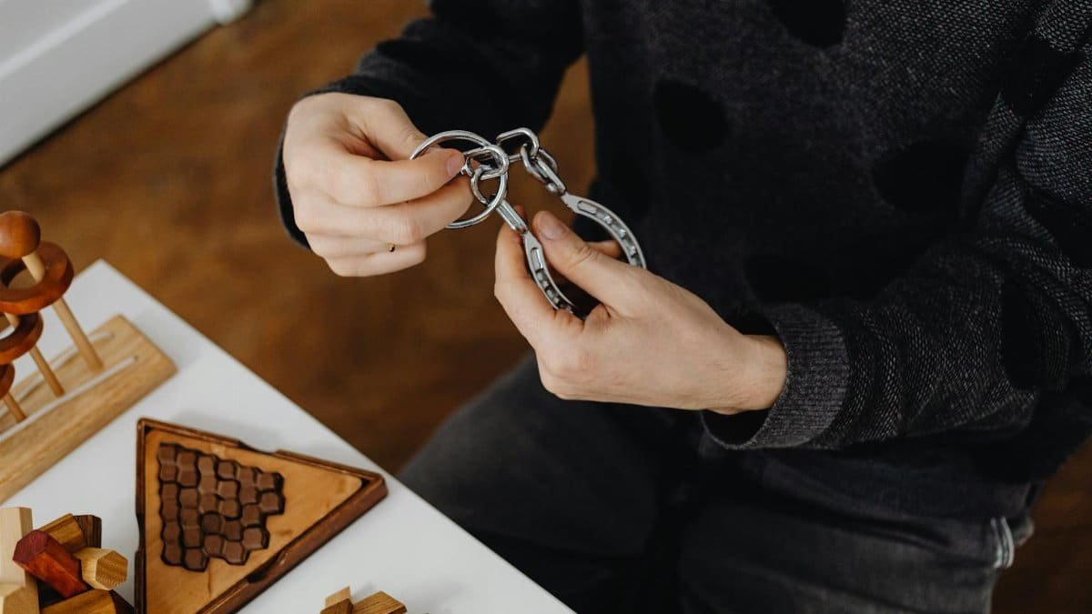 Top view of hands solving metal puzzle, surrounded by wooden brain teasers on a table.