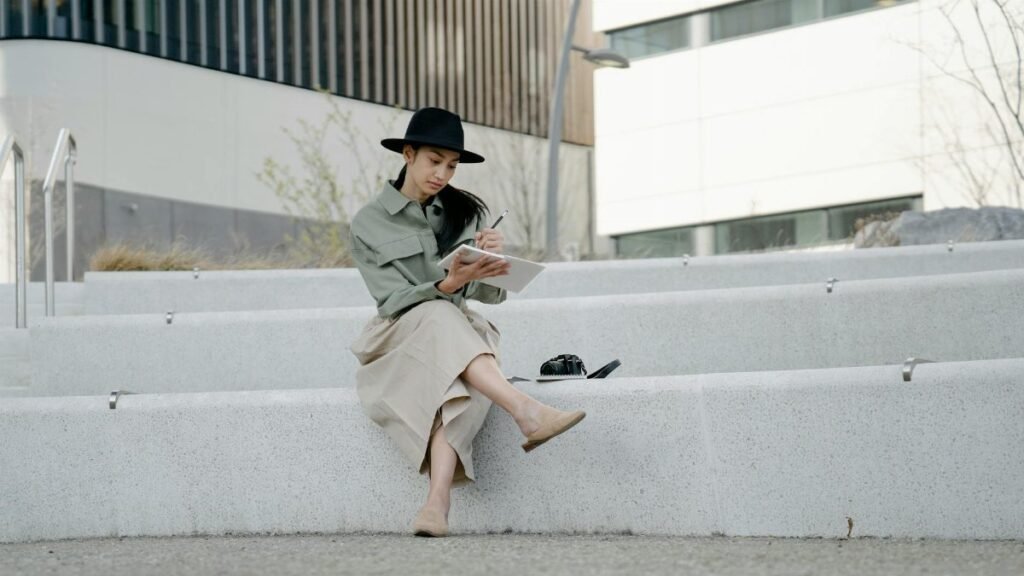 Woman in hat sitting on steps outdoors, writing in a notebook with crossed legs.