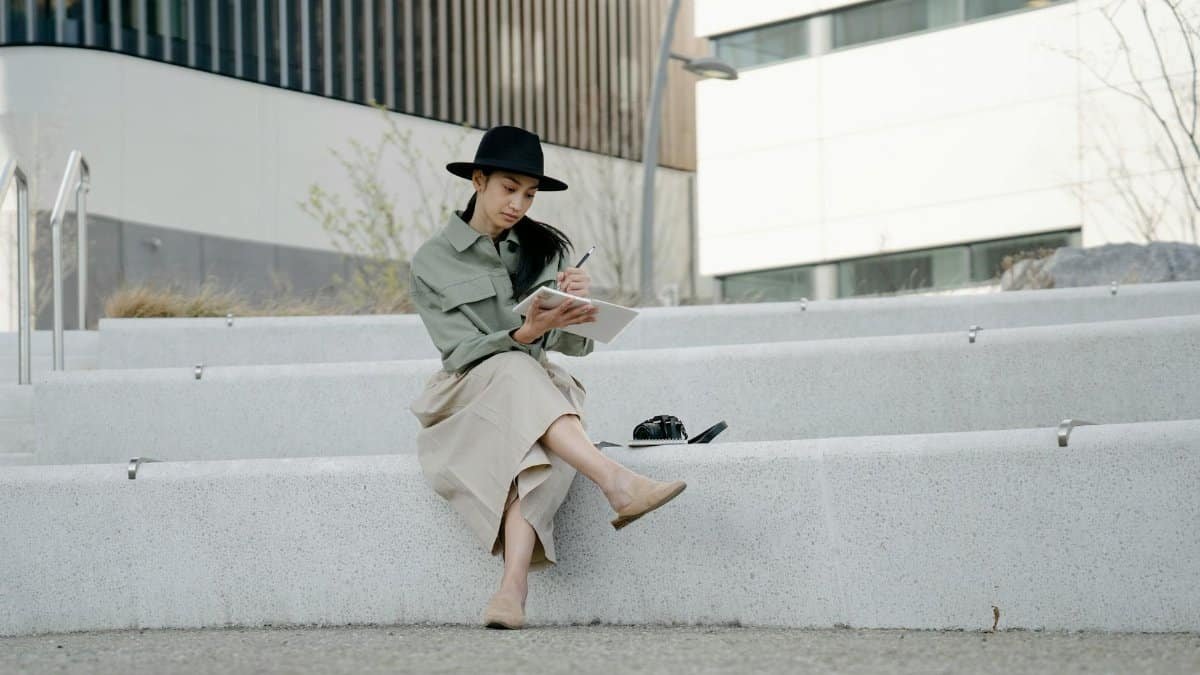 Woman in hat sitting on steps outdoors, writing in a notebook with crossed legs.