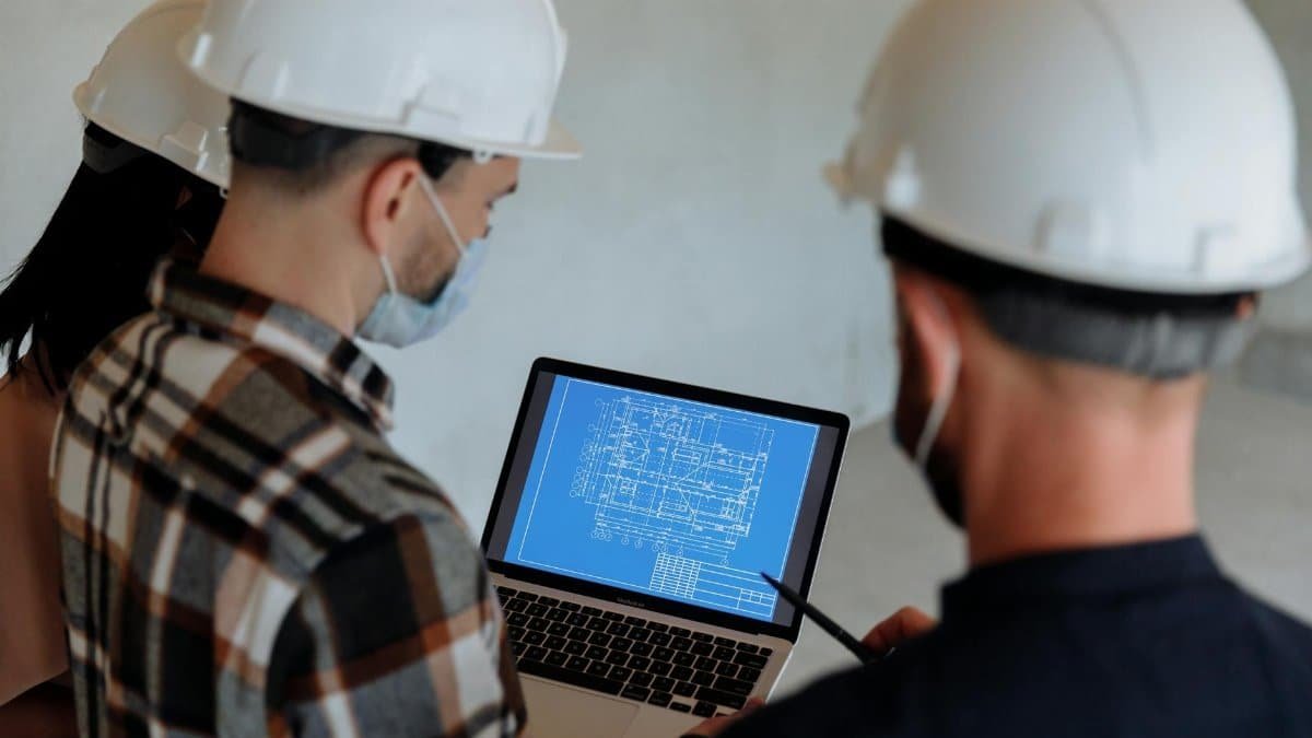 Engineers wearing hard hats and masks review a construction blueprint on a laptop indoors.