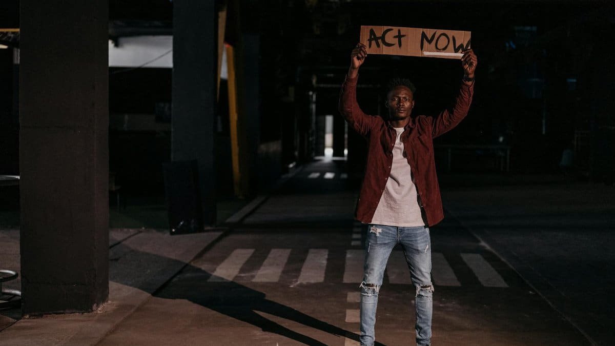 A man stands holding an 'Act Now' sign in an urban environment, emphasizing activism.