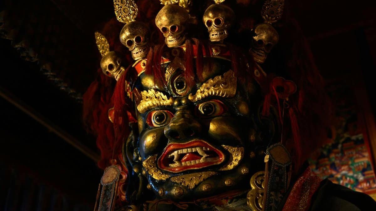From below of Tsam mystery mask with gilded human face and third eye and pig nose and fangs among teeth and golden circles around face and full head of red hair decorated with red hairband adorned with golden skulls