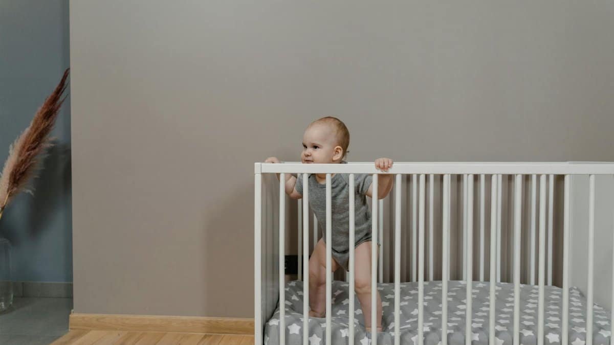 A cute baby in a grey onesie stands in a white crib indoors.