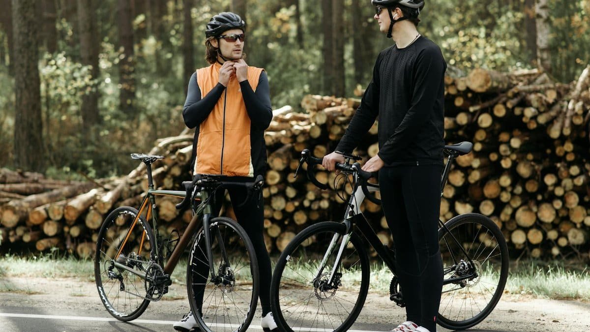 Two male cyclists pause on a forest road beside stacked logs, wearing helmets and athletic gear.