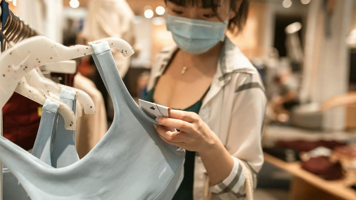 Woman in a store examining clothing tags while wearing a face mask, embracing new normal shopping habits.