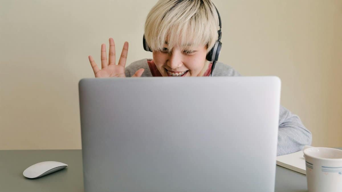 Smiling young Asian female in headphones and casual clothes having conversation via netbook and giving high five while looking at screen and sitting at table with cup of hot drink