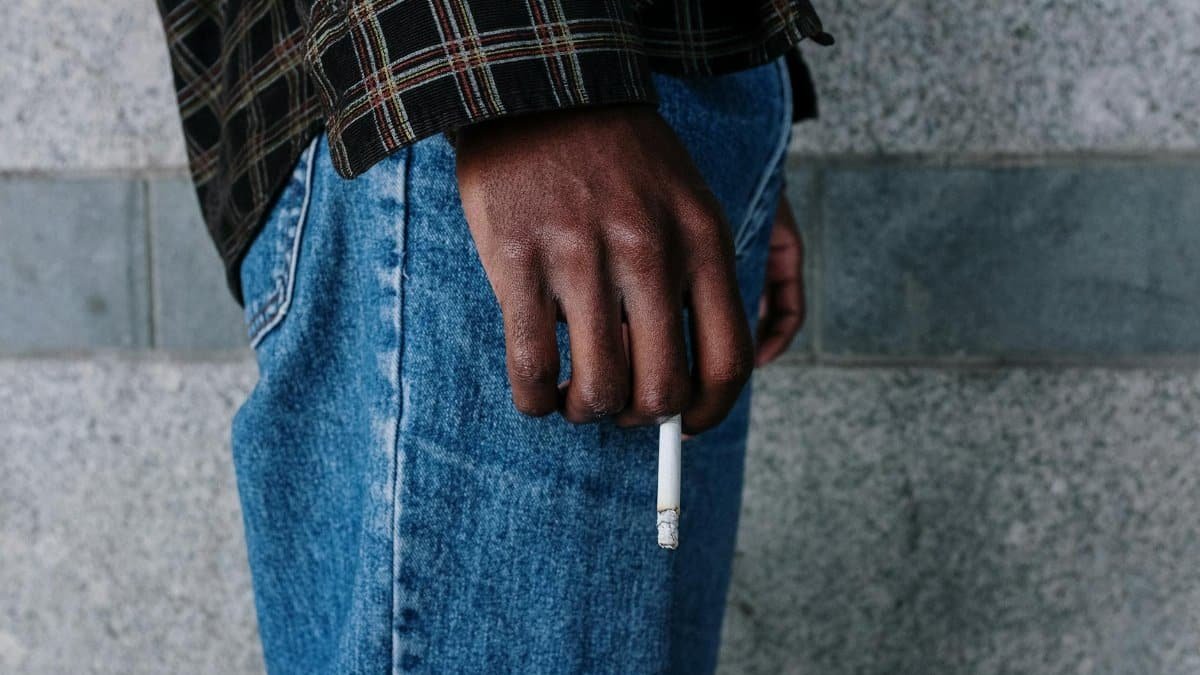 Close-up of a person holding a cigarette against an urban stone background, showcasing casual fashion.