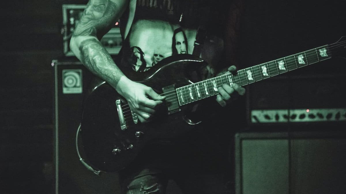 Musician with tattoos playing an electric guitar on stage in a dark, moody setting.