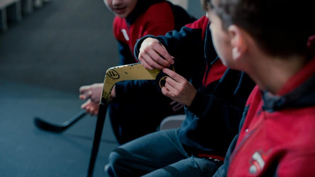 Children in a locker room taping hockey sticks, preparing for a game.