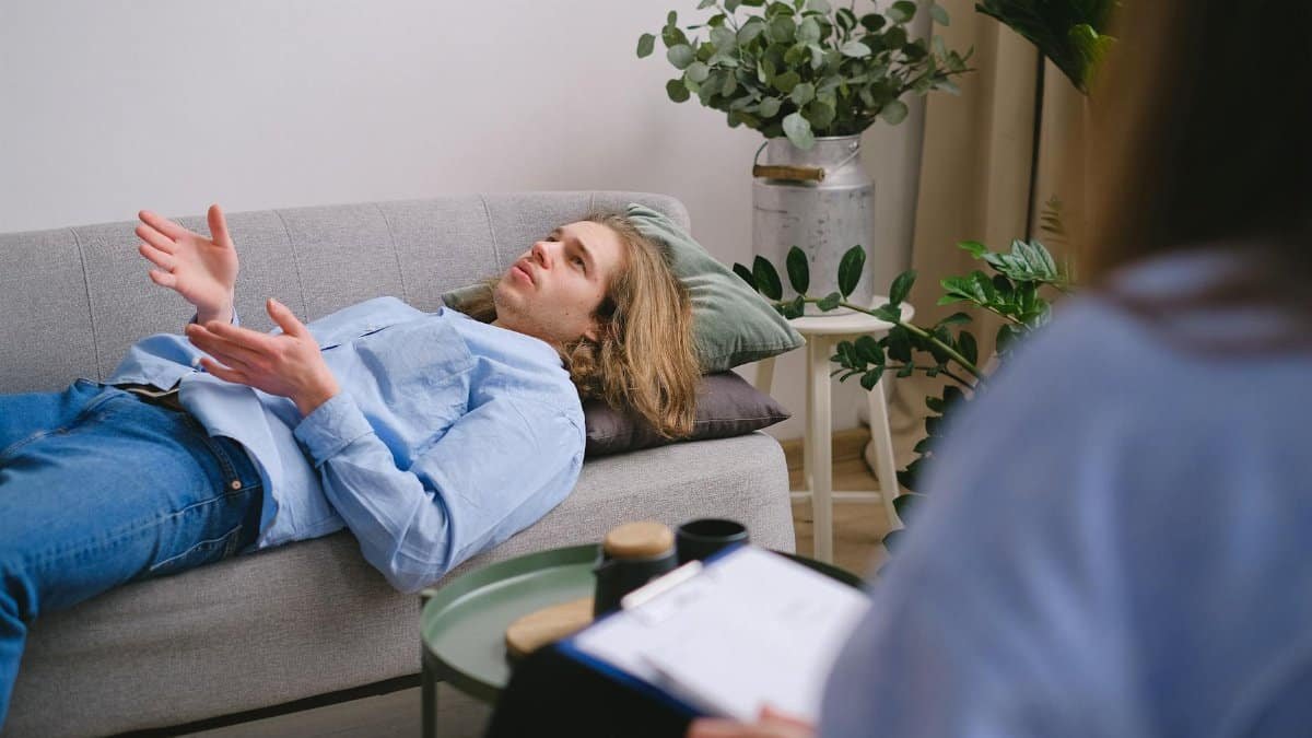 Concerned male client lying on sofa and speaking about mental problems with unrecognizable psychologist with clipboard during psychotherapy appointment in office