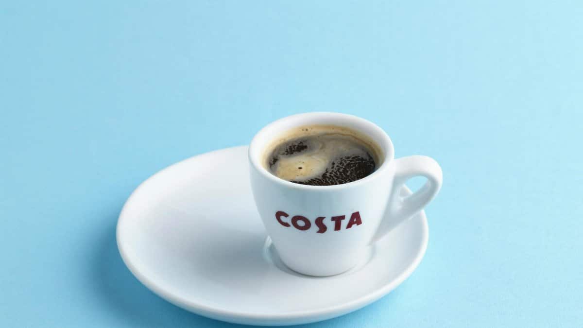 Close-up of a Costa coffee cup and saucer with dark coffee against a light blue backdrop.