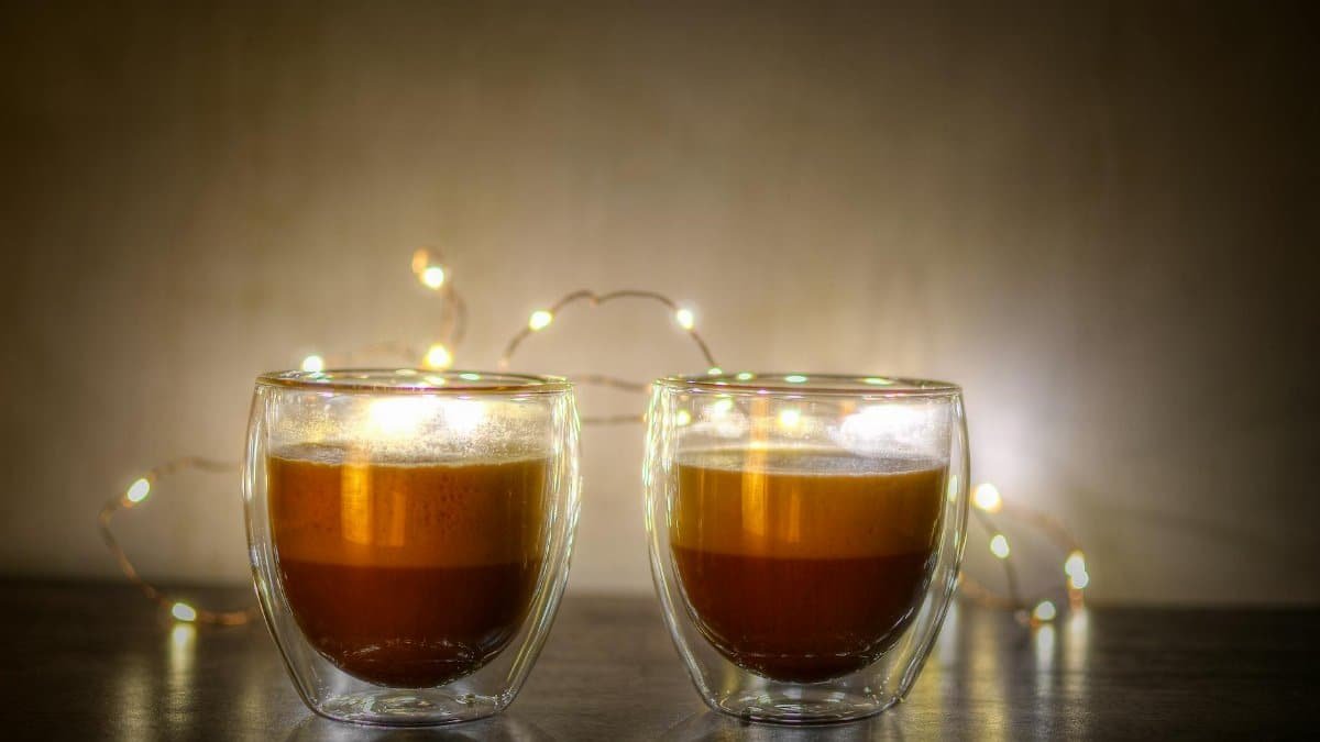 Two cappuccinos in double-walled glasses with festive lights.