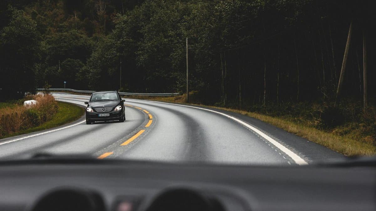 A black car driving on a curved road surrounded by Norwegian forest scenery.