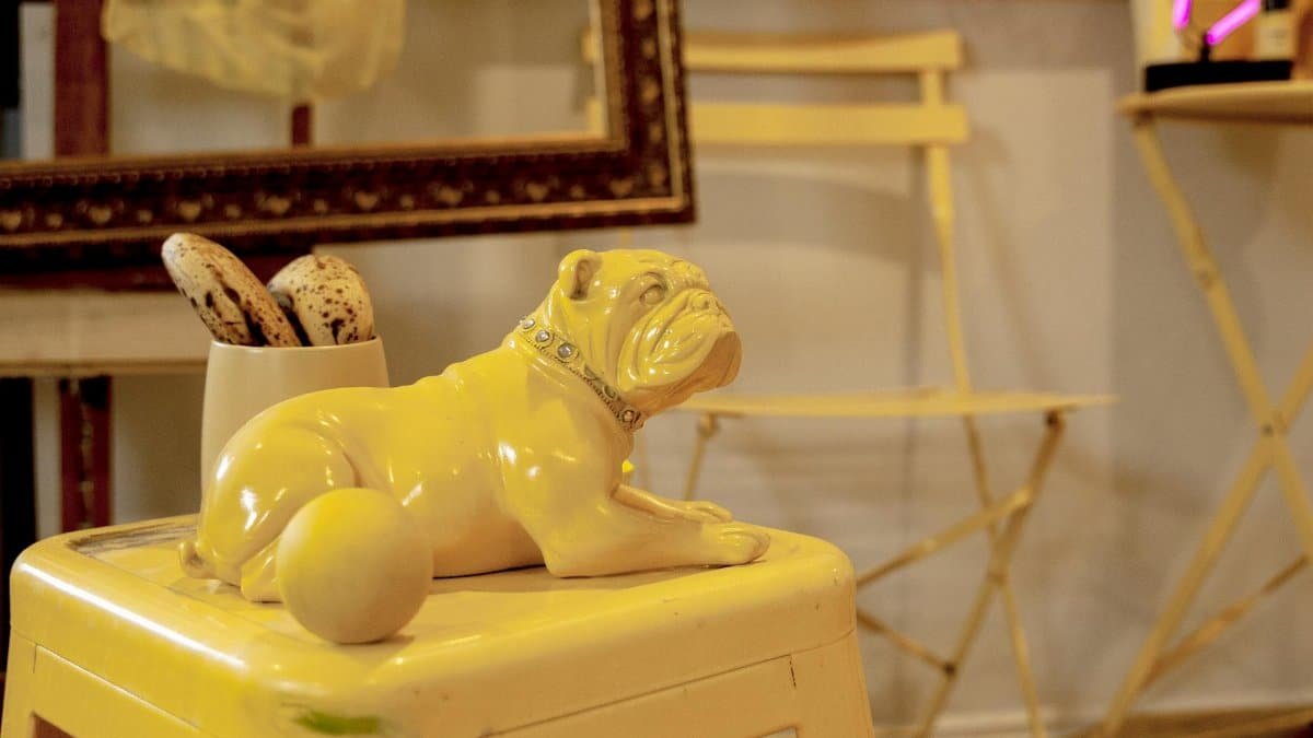 A yellow bulldog figurine sits on a stool in an art studio filled with chairs and decorative items.