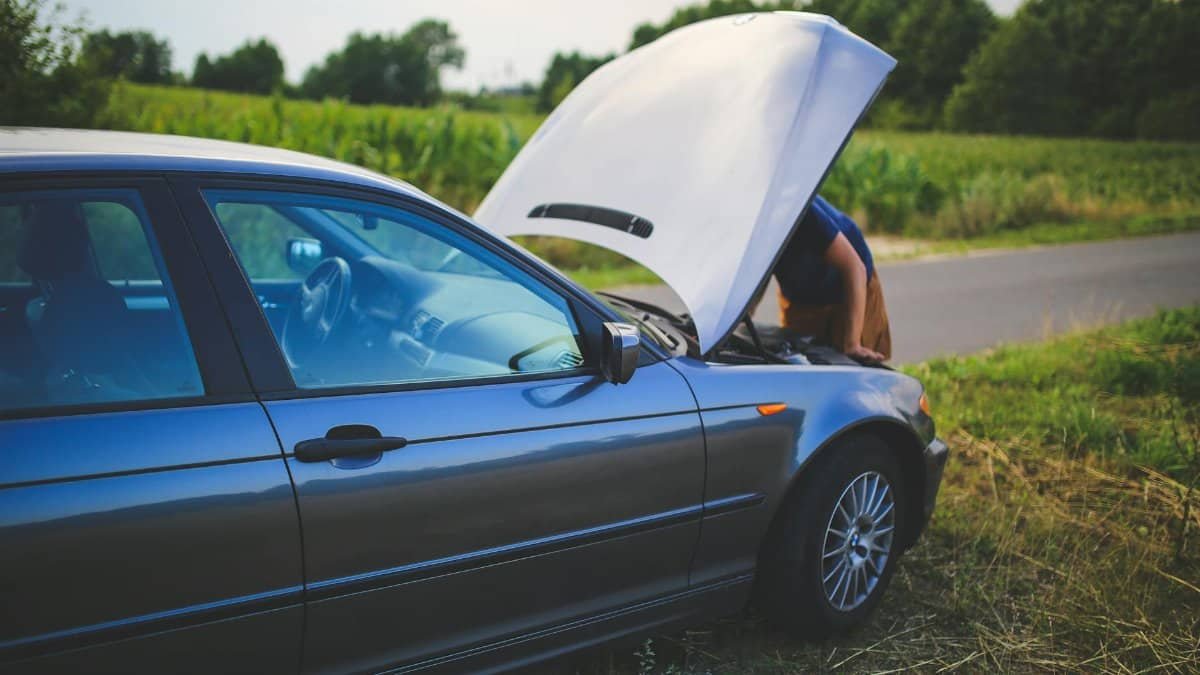 Man checking car engine with hood open by the side of a rural road.