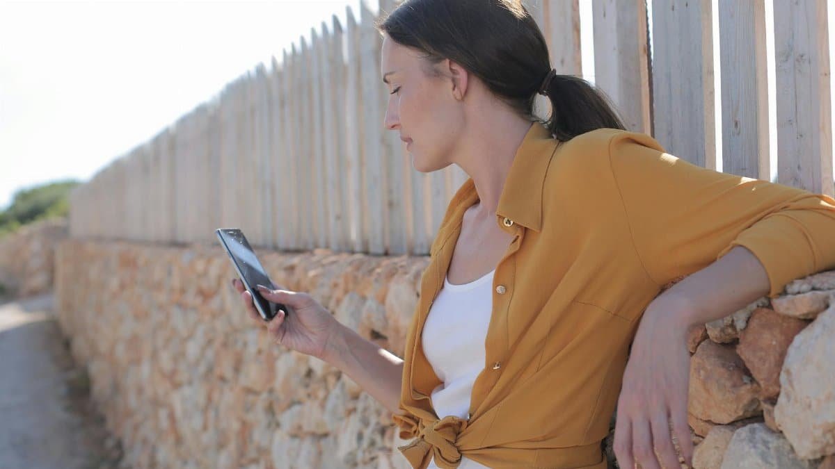 Woman leaning against stone wall using smartphone outdoors on a sunny day.