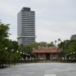 A towering skyscraper stands behind a historic pagoda amid trees in an urban park.