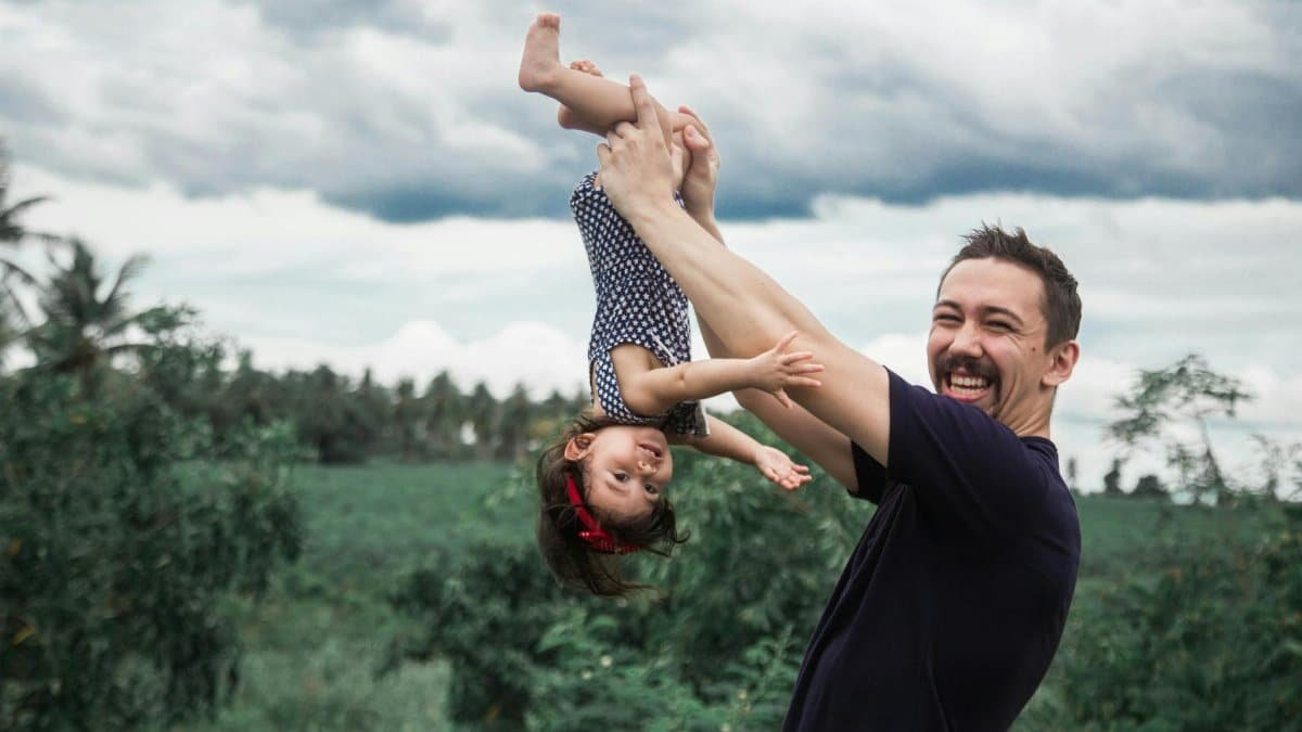 A joyful father lifts his daughter upside down in an outdoor setting.