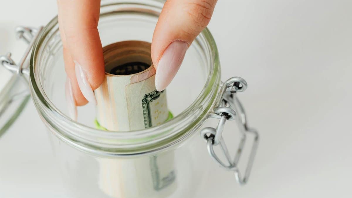 Close-up of hand placing rolled currency into a clear glass jar, symbolizing savings.