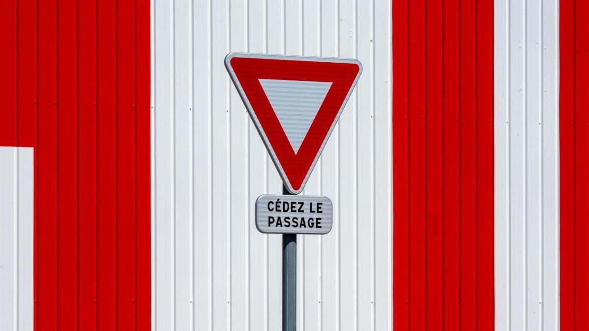 A French 'Yield' sign against a red and white striped wall, minimalistic design.