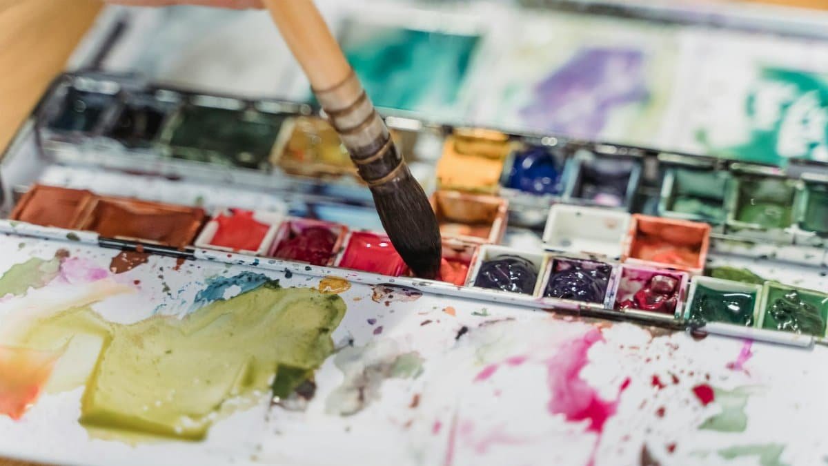 Colorful watercolor palette in use with a paintbrush applying paint, showcasing messy artistic creativity.
