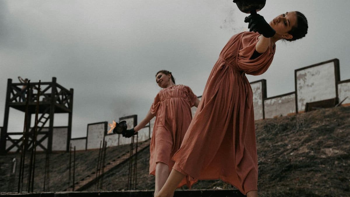 Two women perform an elegant dance with fire, set outdoors against a dramatic backdrop.