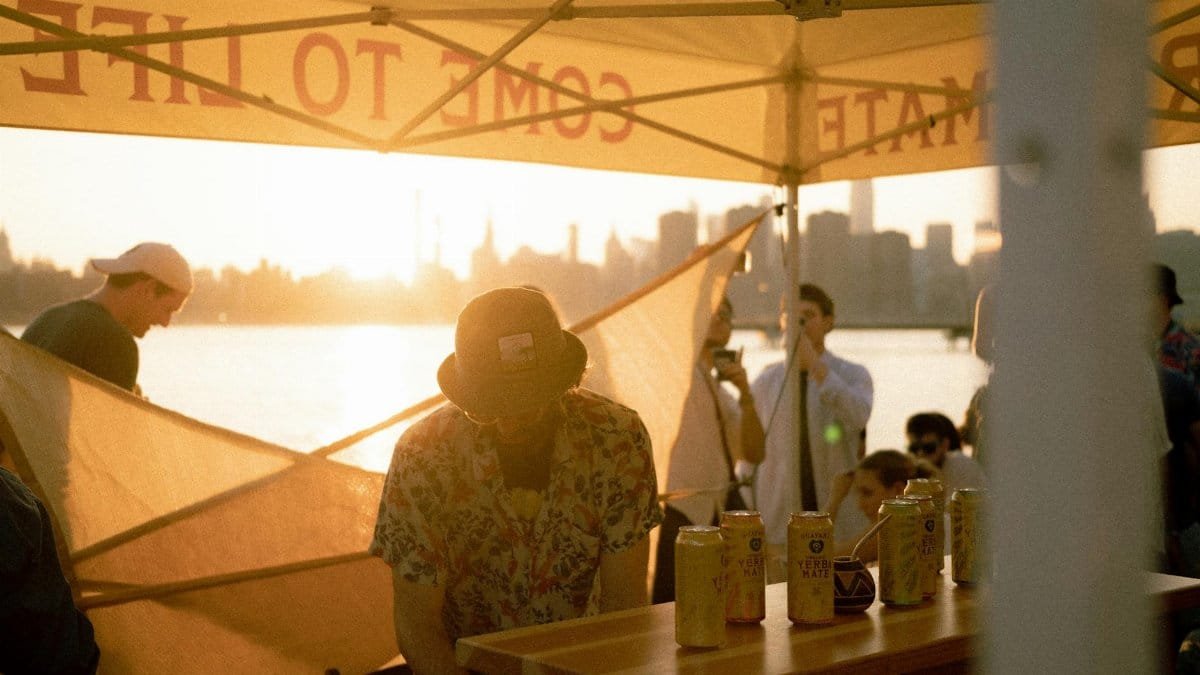People gathering under a tent near waterfront with New York City skyline in the background during sunset.