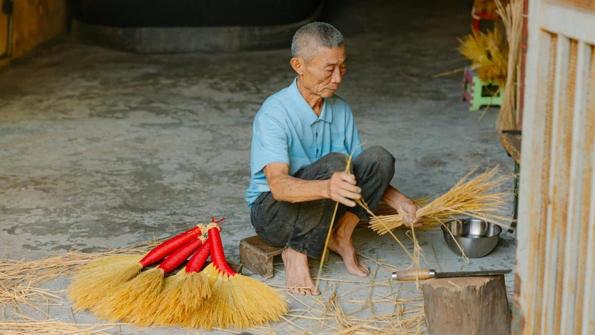 Asian elderly man crafting handmade brooms indoors, showcasing traditional skills.