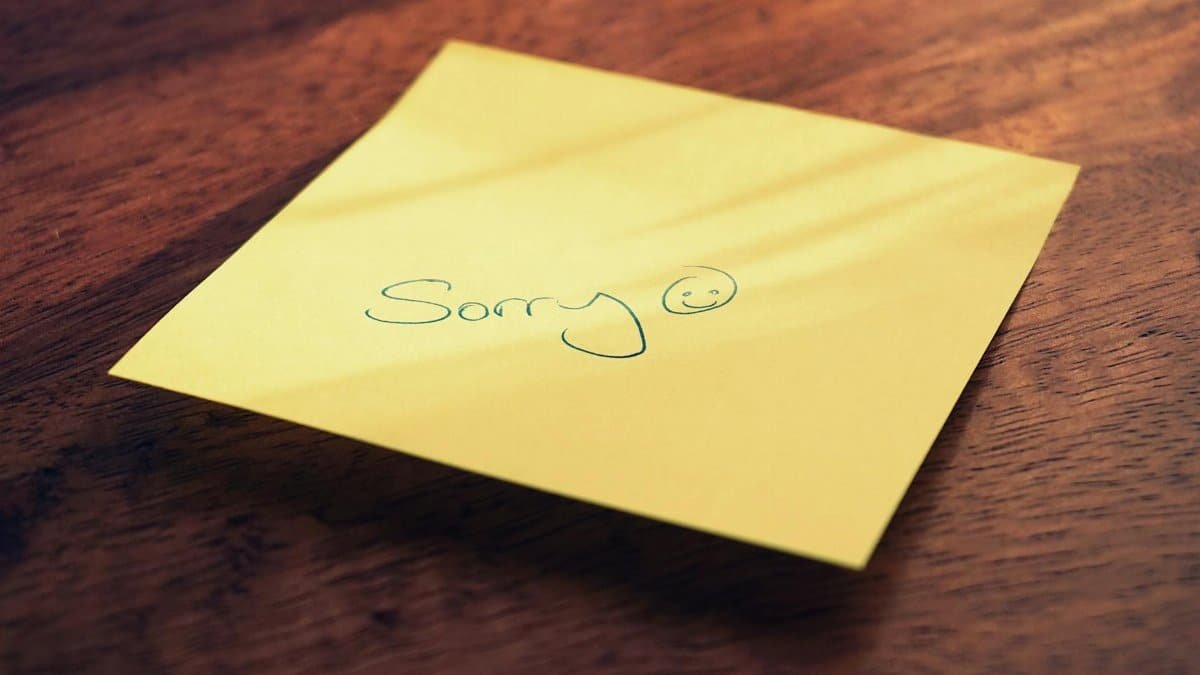 A yellow sticky note with a handwritten apology message and smiley face on a wooden surface.