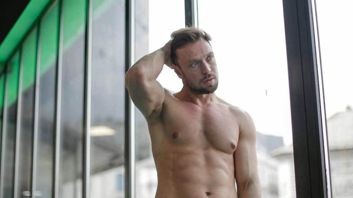 A shirtless muscular man standing indoors by a window, showcasing strength and masculinity.