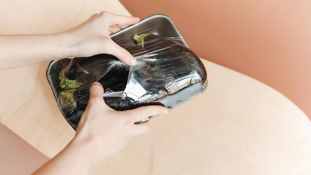 Hands holding a plastic-wrapped tray of eggplants on a wooden surface, emphasizing zero waste.