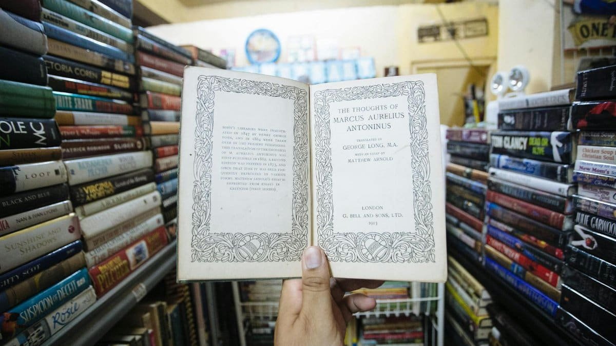 A hand holds an open book with decorative text in a vintage bookstore, surrounded by shelves of books.