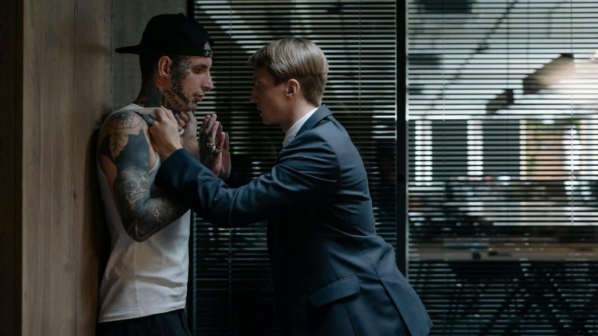 Business-suited detective confronts tattooed suspect in tense indoor setting, emphasizing investigation.