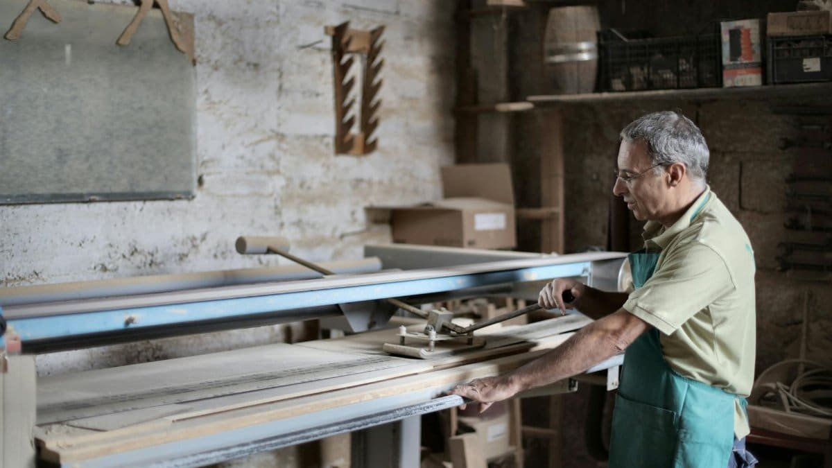Side view of serious craftsman in green apron using manual machine while polishing detail during workday in workshop on sunny day