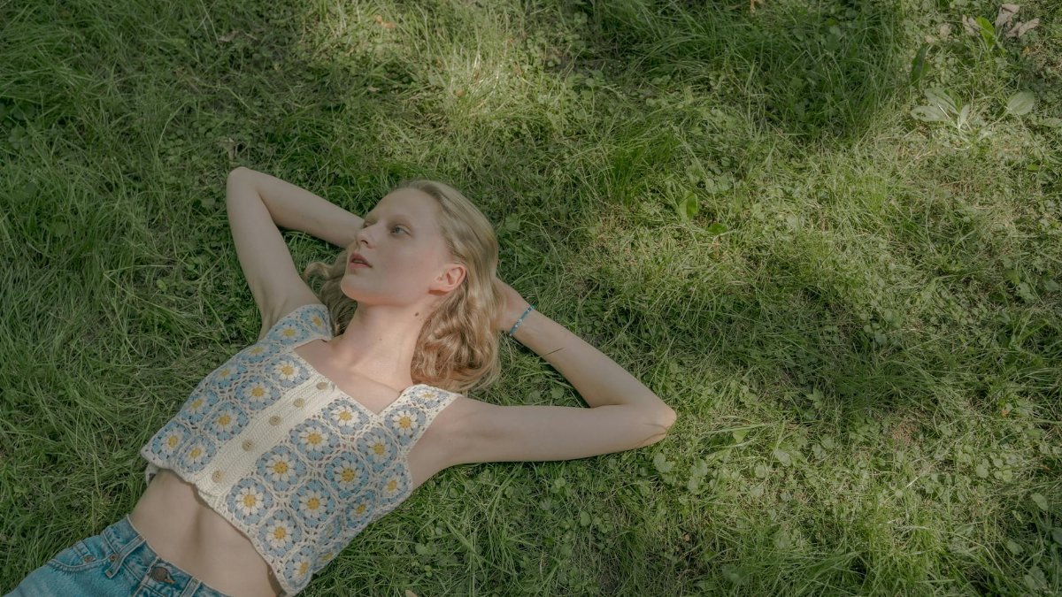 A young woman with blonde hair lays on green grass, enjoying a sunny summer day.