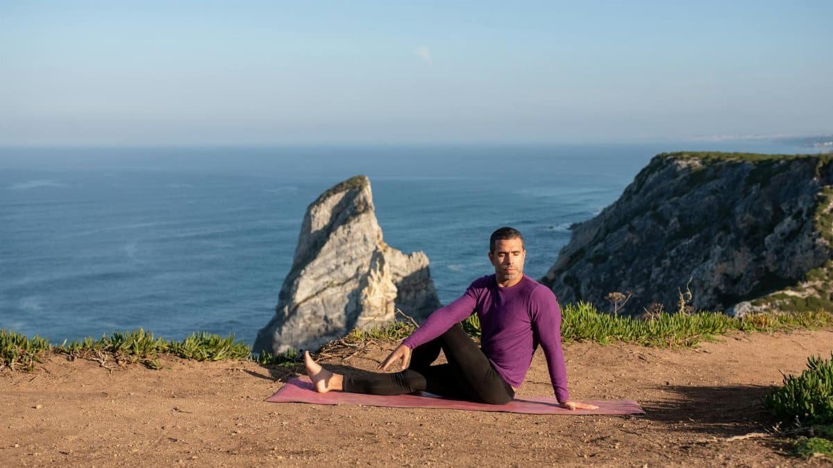 Man practicing yoga seated twist pose on cliffs in Portugal with ocean and cliffs view.