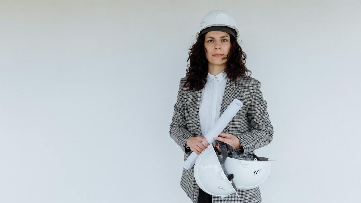 Engineer in formal attire holding blueprints and hard hats, standing against a white wall.