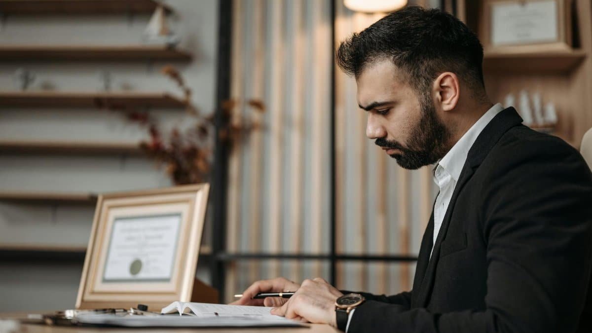 A professionally dressed man focused on writing at a desk within an office setting, suggesting a serious work environment.