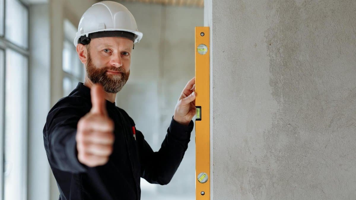 Professional construction worker with hard hat and spirit level giving thumbs up indoors.