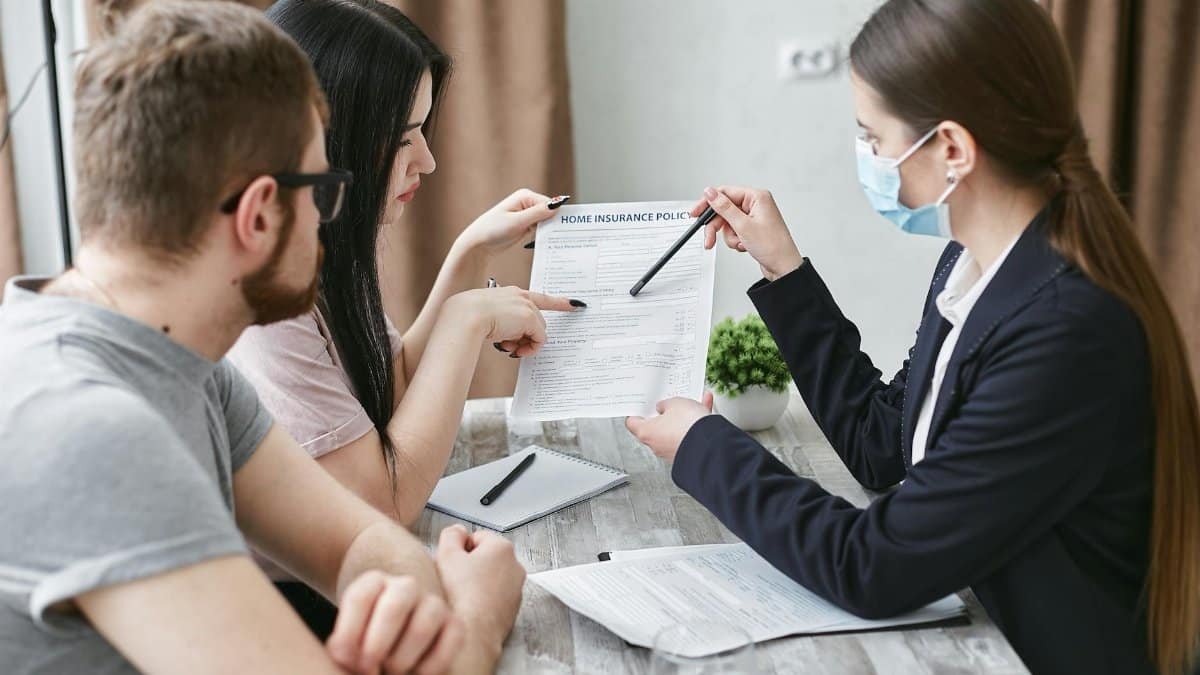 Three adults discuss a home insurance policy at a meeting table indoors.
