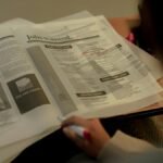 Person reviewing job listings in a newspaper, holding a pink marker for highlighting job ads.