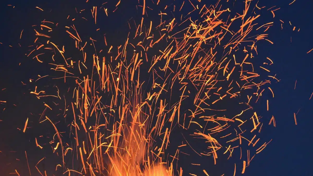 Vibrant orange sparks fly from a fire against a dark blue night sky, creating a dramatic visual.
