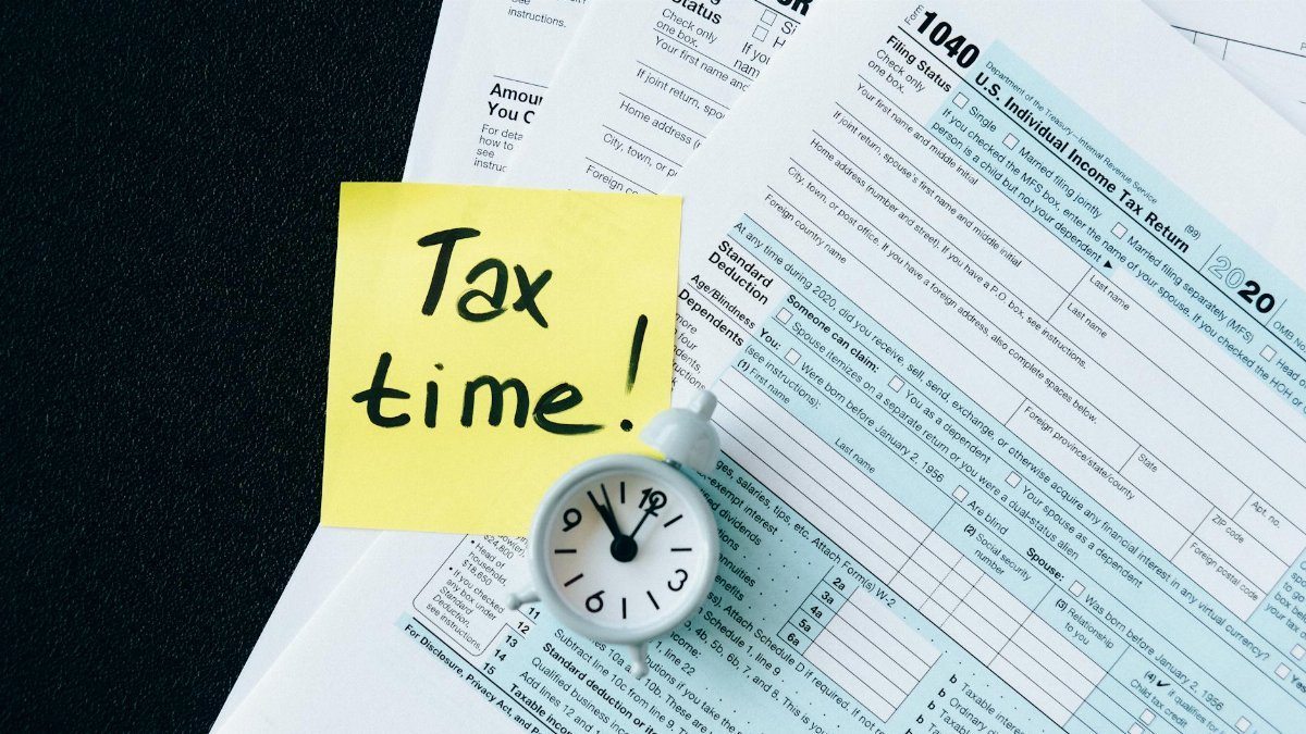 A stack of tax forms with a clock and yellow sticky note saying 'Tax time!' indicating urgency.